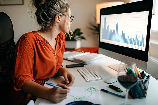 Frau sitzt am Schreibtisch und schaut auf einen Monitor mit einem Diagramm.