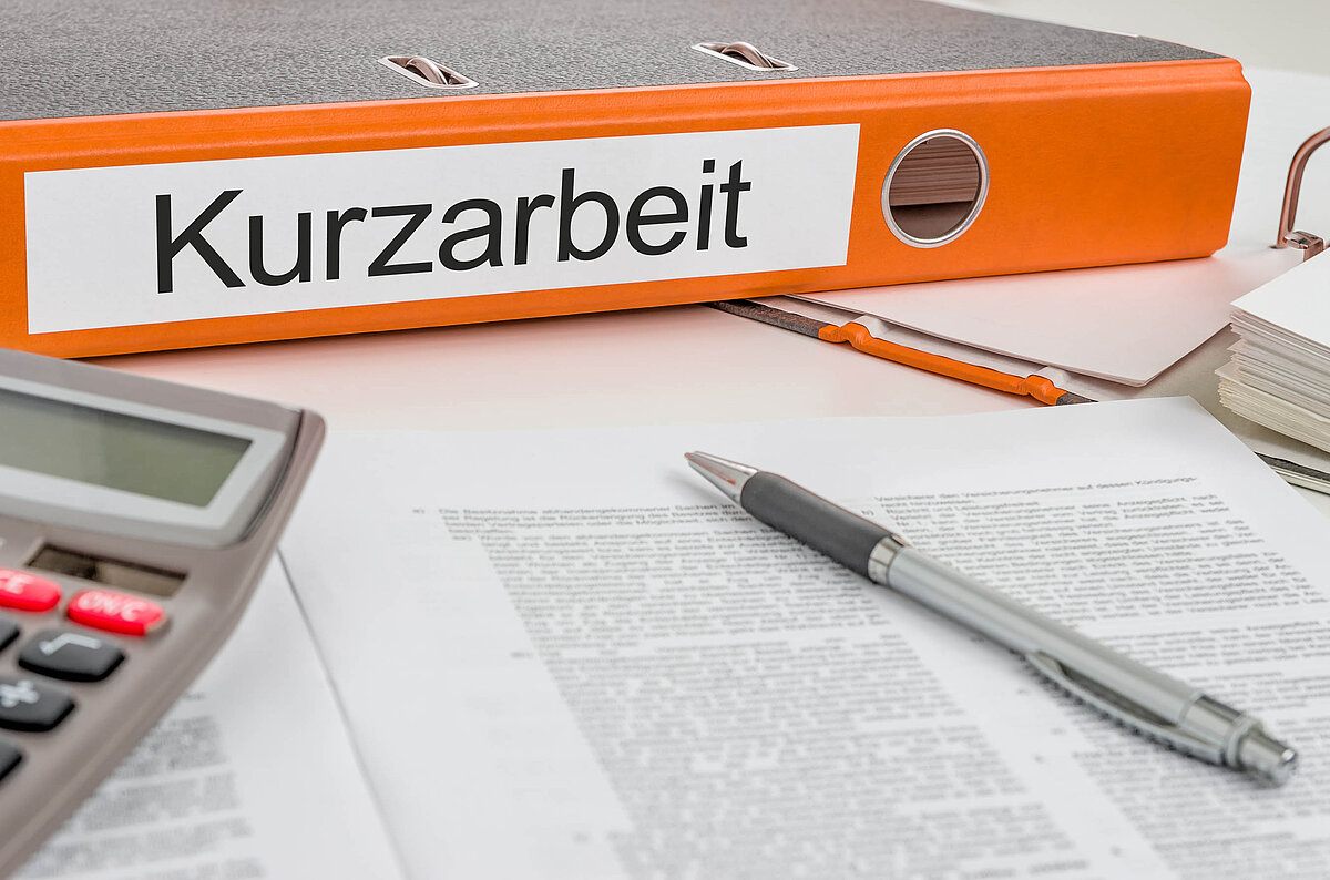 Auf einem Schreibtisch mit Papieren und einem Kugelschreiber liegt ein Ordner mit der Aufschrift "Kurzarbeit"