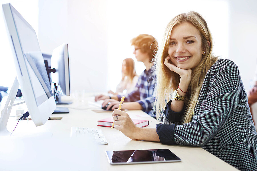 Mehrere fröhliche Personen sitzen vor iMac-Computern in einem Schulungsraum.
