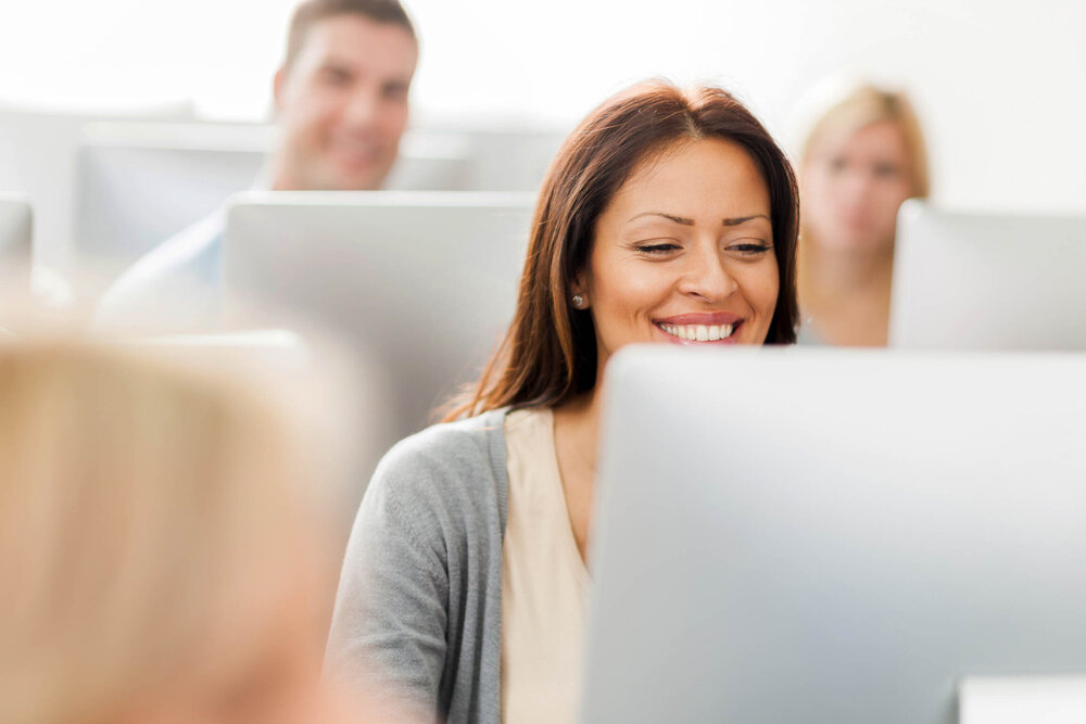 Mehrere fröhliche Personen sitzen vor iMac-Computern in einem Schulungsraum.