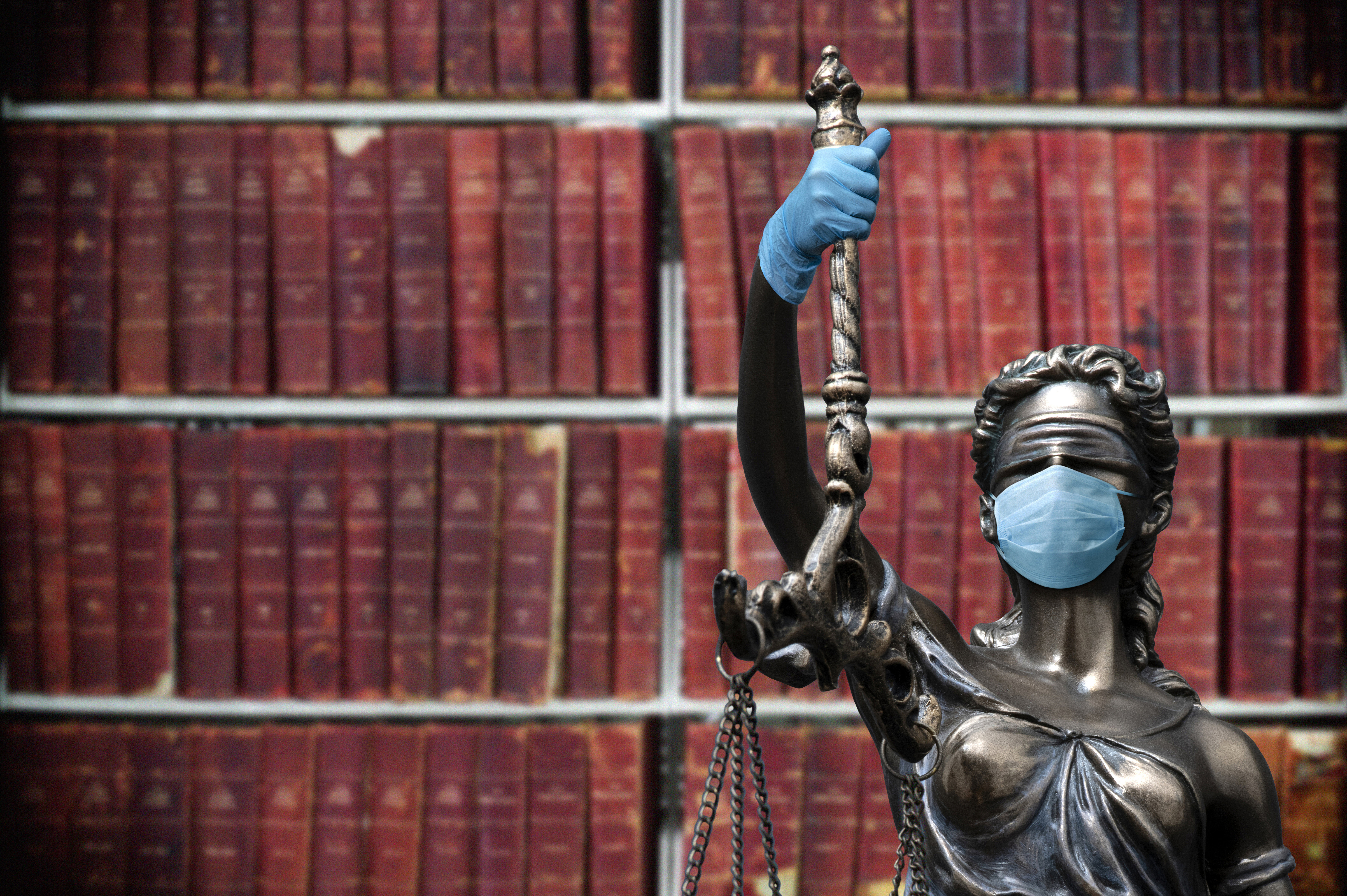 Human Rights Law in the Time of the Coronavirus Eine Justizia-Figur aus Metall steht vor einem Bücherregal und trägt Gummihandschuhe sowie eine Atemschutzmaske.