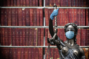 Eine Justizia-Figur aus Metall steht vor einem Bücherregal und trägt Gummihandschuhe sowie eine Atemschutzmaske.